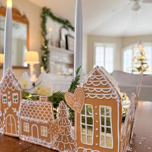 Könnte beinhalten: Ein Lebkuchenhaus-Mittelstück mit weißem Zuckerguss und einem Herzdetail. Die Häuser sind in einer Reihe angeordnet, mit einem kleinen immergrünen Baum in der Mitte. Das Mittelstück wird mit weißen Kerzen beleuchtet.