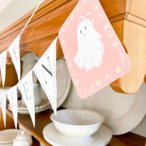 May include: A white and black Halloween banner with the word "Happy" and a ghost illustration. The banner is hanging on a wooden shelf with white dishes.