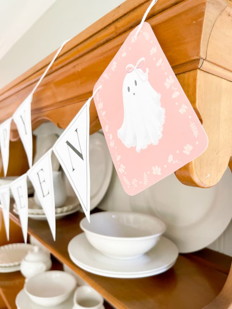 May include: A white and black Halloween banner with the word "Happy" and a ghost illustration. The banner is hanging on a wooden shelf with white dishes.