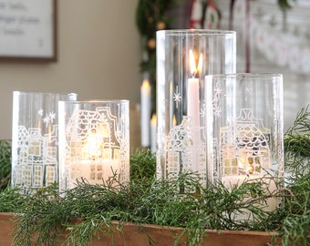 May include: Four clear glass candle holders with white snowflake designs and lit candles are arranged in a wooden bowl filled with green foliage.