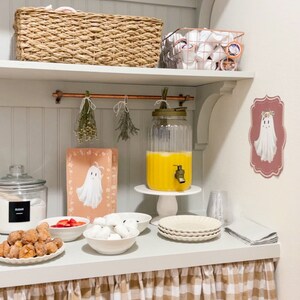 May include: A white shelf with a brown wicker basket, a copper pipe, and a glass dispenser filled with orange juice. There are also white plates, bowls, and a ghost-themed sign on the wall. The shelf is decorated with a brown and white checkered tablecloth.