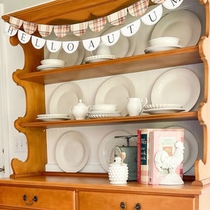 May include: A wooden hutch with two drawers and shelves. The hutch is decorated with a banner that reads "Hello Autumn" and has a plaid pattern. The shelves are filled with white dishes and a rooster figurine.