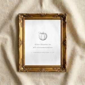 May include: A rectangular wall art print in an ornate gold frame. The white print features a black line drawing of a pumpkin centered above the text: Give thanks in all circumstances, 1 THESSALONIANS 5:18, printed in a simple black serif font.