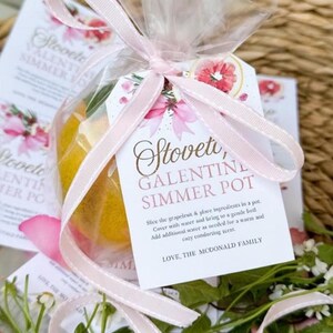 Könnte beinhalten: Ein Geschenkset mit Grapefruit und Grünzeug, verpackt in durchsichtigem Zellophan und mit einem rosa Band gebunden. Das Etikett lautet "Stoveti, Galentine Simmer Pot" mit Anweisungen und einem floralen Design.