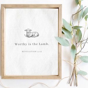 Op de afbeelding: Een ingelijste print met een licht houten frame. De print toont een zwart-wit illustratie van een lam en de tekst "Worthy is the Lamb. Openbaring 5:12". Een bundel eucalyptus takken vastgebonden met touw staat naast het frame.