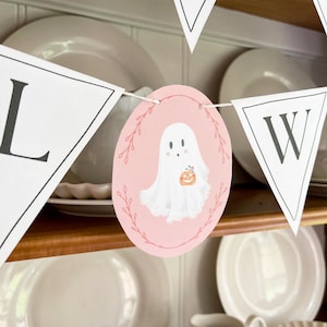 May include: A pink and white Halloween banner with the word "Halloweeen" spelled out in black letters. The banner features a ghost holding a pumpkin.