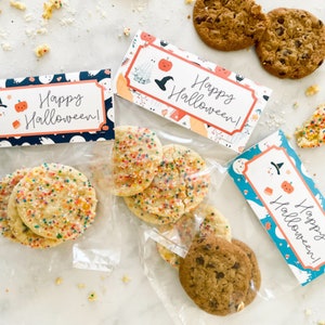 May include: Three clear plastic bags filled with round cookies, each sealed with a folded paper topper featuring the text Happy Halloween and illustrations of pumpkins, ghosts, and witch hats. Some cookies have colorful sprinkles, others chocolate chips.
