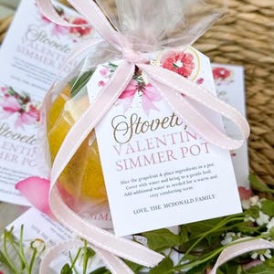 Peut inclure: Un ruban rose noué autour d'un sac en cellophane transparent rempli d'un pamplemousse et de brins de romarin. Une étiquette blanche avec des accents floraux roses indique "Stoveti, Valentin Simmer Pot" et comprend des instructions pour faire un pot mijoté. L'étiquette comprend également le texte "LOVE, THE MCDONALD FAMILY".