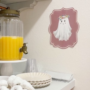May include: A white ghost with a floral crown on a pink background. The ghost is wearing a white dress with lace trim. The background is a pink rectangle with a scalloped edge. The image is a cute and whimsical illustration of a ghost.