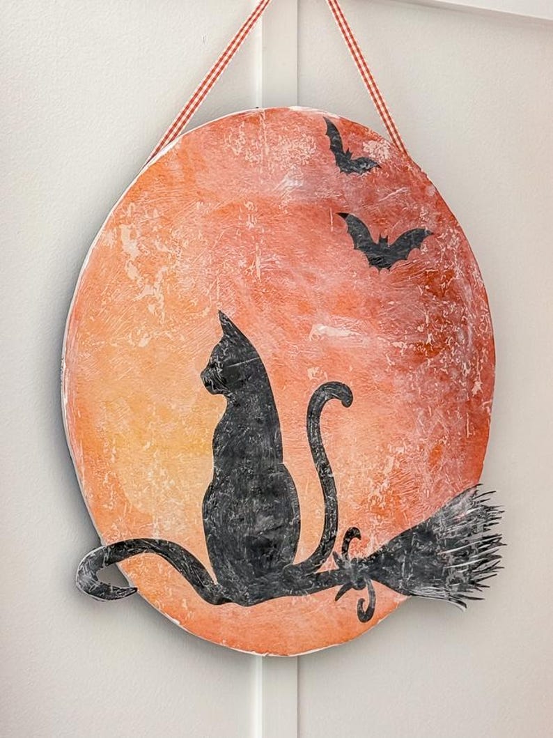 Close-up of a handmade black cat Halloween door hanger with an orange moon, bats and a distressed vintage finish, hung with gingham ribbon