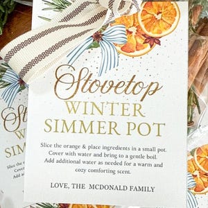 Pode incluir: Uma etiqueta de presente para uma panela de inverno para fogão, com as palavras "Stovetop Winter Simmer Pot" em escrita dourada. A etiqueta é decorada com fatias de laranja, laços azuis e uma fita listrada.