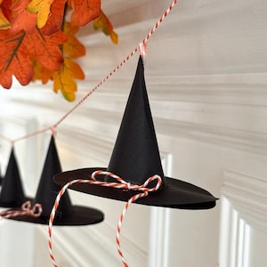 Puede incluir: Guirnalda de sombreros de bruja negros con hilo rojo y blanco, suspendidos frente a una pared blanca. Los sombreros están decorados con un lazo rojo y blanco. Hojas de otoño en tonos naranja y amarillo en el fondo.