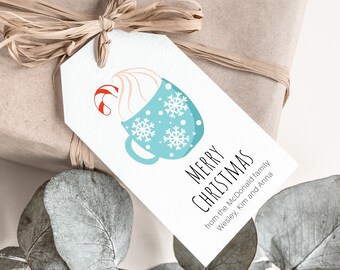 May include: A white gift tag with a blue mug design, snowflakes, and a candy cane. The tag reads "Merry Christmas" and includes a family name. The tag is tied to a wrapped gift with raffia.