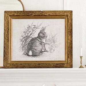 May include: A black and white illustration of a rabbit sitting in a field of tall grass, framed in a gold ornate frame.