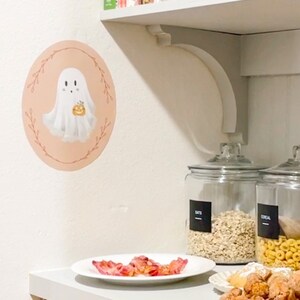May include: A white ghost holding a small orange pumpkin, with a pink background and a floral wreath, is on a white wall. A white shelf with a white ceramic rooster and a copper wire basket is above the ghost. A white shelf below the ghost has a plate of bacon, a glass jar labeled "OATS", a glass jar labeled "CEREAL", and a plate of fried dough balls.