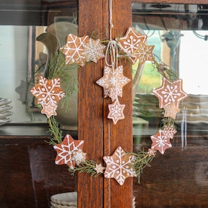 May include: A wreath made of greenery with gingerbread snowflake ornaments. The wreath is hanging on a wooden cabinet door.