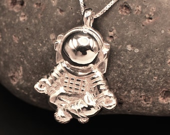 May include: Silver pendant necklace featuring a detailed astronaut charm. The astronaut is wearing a spacesuit with a helmet and is holding a small object in its hand. The necklace is on a silver chain and is resting on a grey rock. The text "Resurgence. Silver Studio Los Angeles" is visible in the top right corner of the image. The text "MADE IN USA" is visible in the bottom right corner of the image.