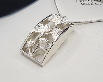 May include: A silver pendant necklace featuring a delicate design of ginkgo leaves. The pendant is shaped like a rectangle with rounded edges and has a cutout design of the leaves. The necklace is on a silver chain. The pendant is made in the USA. "Resurgence Art Studio Los Angeles" is printed on the image.