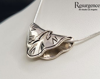 May include: A silver necklace featuring a distinctive pendant shaped like a butterfly with Roman numeral markings. The pendant is attached to a silver chain. The necklace is made in the USA. The text "Resurgence Art Studio Los Angeles" is visible in the image.