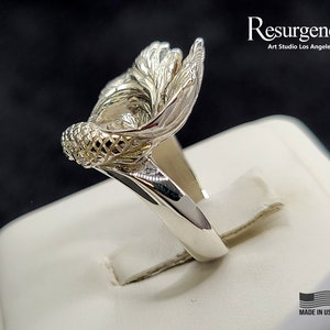 May include: A silver ring featuring an intricate fish tail design. The ring is displayed on a white stand. The text "Resurgence. Art Studio Los Angeles" and "Made in USA" are visible on the stand.