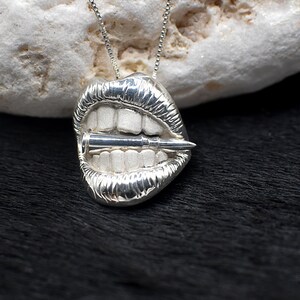 May include: Silver pendant necklace featuring a detailed depiction of a mouth with a bullet in it.