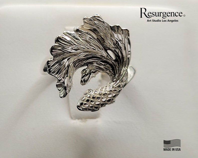 May include: A silver ring with a detailed, three-dimensional design of a betta fish with flowing fins. The ring is made in the USA. "Resurgence Art Studio Los Angeles" is printed on the white background.