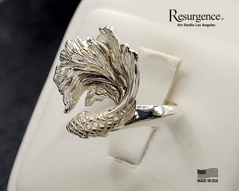 May include: A silver ring featuring a detailed, intricate design of a fish with flowing fins. The ring is made in the USA.