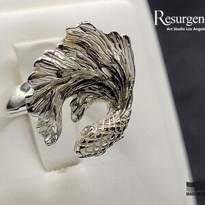 May include: A silver ring featuring a detailed, three-dimensional design of a betta fish with flowing fins. The ring is made by Resurgence Art Studio Los Angeles and is marked "Made in USA".