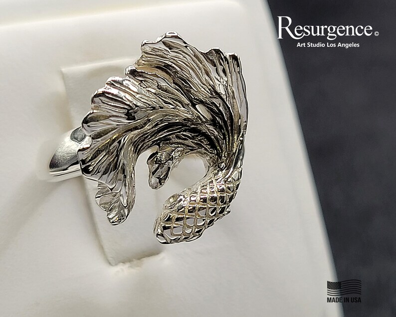 May include: A silver ring featuring a detailed, three-dimensional design of a betta fish with flowing fins. The ring is made by Resurgence Art Studio Los Angeles and is marked "Made in USA".