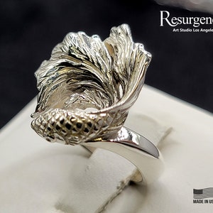 May include: A silver ring featuring a detailed, three-dimensional design of a fish with flowing fins. The ring is made in the USA.