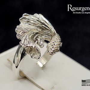 May include: A silver ring featuring a detailed, three-dimensional design of a betta fish. The fish is depicted with flowing fins and a textured body. The ring is made in the USA.