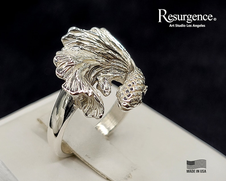 May include: A silver ring featuring a detailed, three-dimensional design of a betta fish. The fish is depicted with flowing fins and a textured body. The ring is made in the USA.