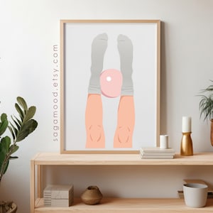May include: A framed art print featuring a minimalist illustration of legs and feet in gray socks, holding a pink object. The artwork is displayed on a light wooden shelf with plants and decorative items. The website sagamood.etsy.com is printed on the side of the frame.