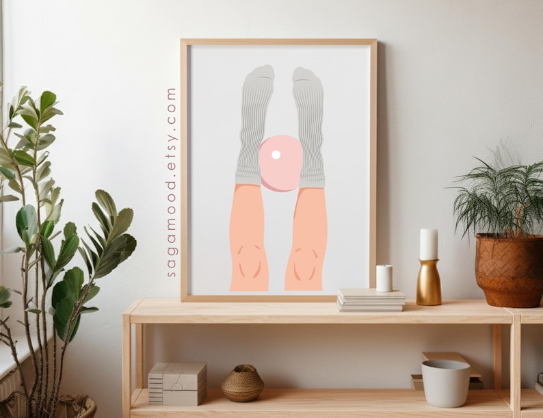 May include: A framed art print featuring a minimalist illustration of legs and feet in gray socks, holding a pink object. The artwork is displayed on a light wooden shelf with plants and decorative items. The website sagamood.etsy.com is printed on the side of the frame.