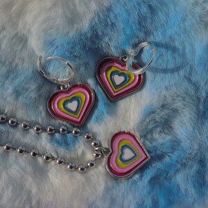 May include: A silver chain necklace with a heart pendant. The pendant is made of metal and has a multi-coloured design with concentric circles of pink, yellow, blue, and white.