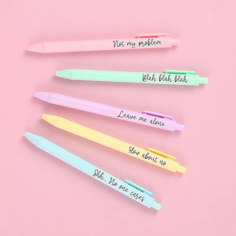 Op de afbeelding: Vijf pastelkleurige pennen met humoristische zinnen erop geschreven. De zinnen zijn "Not my problem", "Blah blah blah", "Leave me alone", "How about no" en "Shh... No one cares".