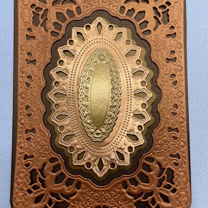 May include: A rectangular, layered die-cut card with an ornate, embossed design. The card features a copper-colored outer frame with floral and scrollwork patterns. Inside, there are olive green and gold oval shapes.