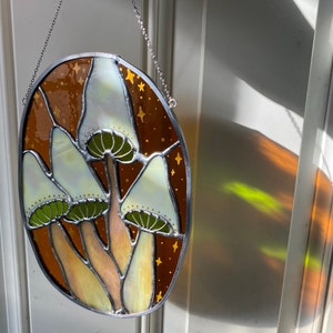 May include: A stained glass window hanging from a chain. The window is oval shaped and features a design of three white mushrooms with green stems and brown caps against a brown background with gold stars.