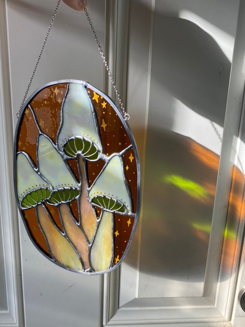 May include: A stained glass window hanging from a chain. The window is oval shaped and features a design of three white mushrooms with green stems and brown caps against a brown background with gold stars.