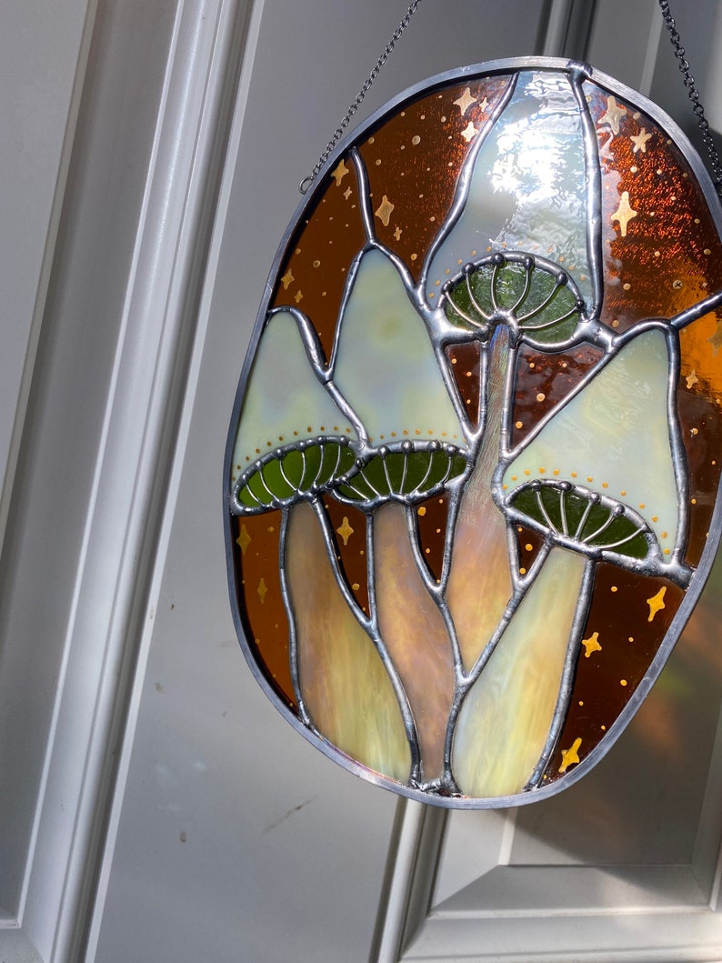 May include: A stained glass window featuring three white mushrooms with green caps and brown stems, set against a brown background with gold stars.