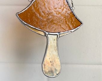 以下が含まれることがあります： 茶色の帽子とクリーム色の茎を持つキノコの形をしたステンドグラスのサンキャッチャー。サンキャッチャーは銀色のチェーンで吊り下げられています。
