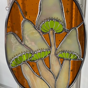 May include: A stained glass window hanging from a chain, featuring four white mushrooms with green caps against a brown background with gold stars.