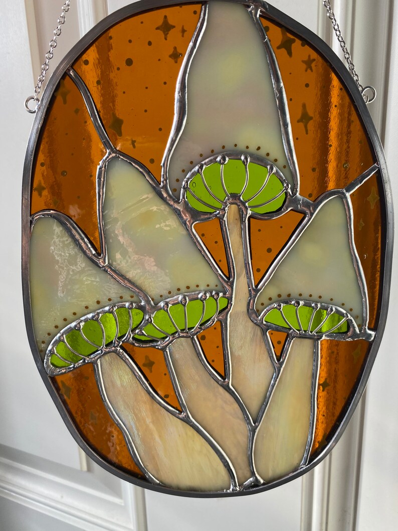 May include: A stained glass window hanging from a chain, featuring four white mushrooms with green caps against a brown background with gold stars.