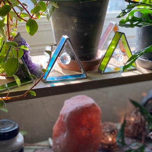 May include: Three glass triangle prism suncatchers with crystal prisms hanging from them. The suncatchers are green, blue, and orange. They are sitting on a wooden shelf with a potted plant in the background.
