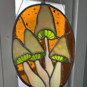 May include: A stained glass window hanging from a chain, featuring three white mushrooms with green caps against a brown background with gold flecks.
