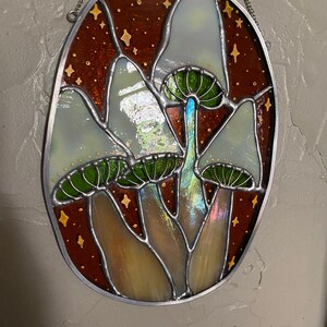 May include: A stained glass window hanging from a chain, depicting three white mushrooms with green caps against a brown background with gold stars.