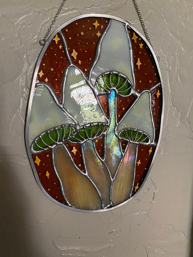 May include: A stained glass window hanging from a chain, depicting three white mushrooms with green caps against a brown background with gold stars.
