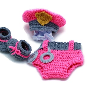 Peut inclure: Une tenue de bébé en crochet rose et gris, comprenant un chapeau avec un badge doré, une couche et des chaussons.