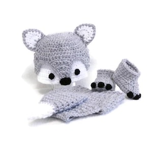 May include: A gray crocheted fox costume for a baby, including a hat, pants, and booties. The hat has fox ears and a black nose, and the booties have black paw prints.