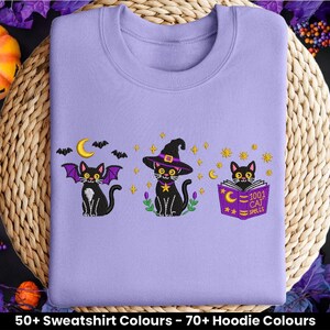 May include: A lilac sweatshirt with embroidered black cats in Halloween designs. One cat has bat wings, another a witch's hat, and the third reads a spell book. The book reads "1001 Cat Spells".